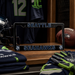 A black Seattle Seahawks license plate frame displayed on a wooden shelf in a locker room setting, surrounded by Seahawks gear including a helmet, folded jerseys, gloves, and a football.