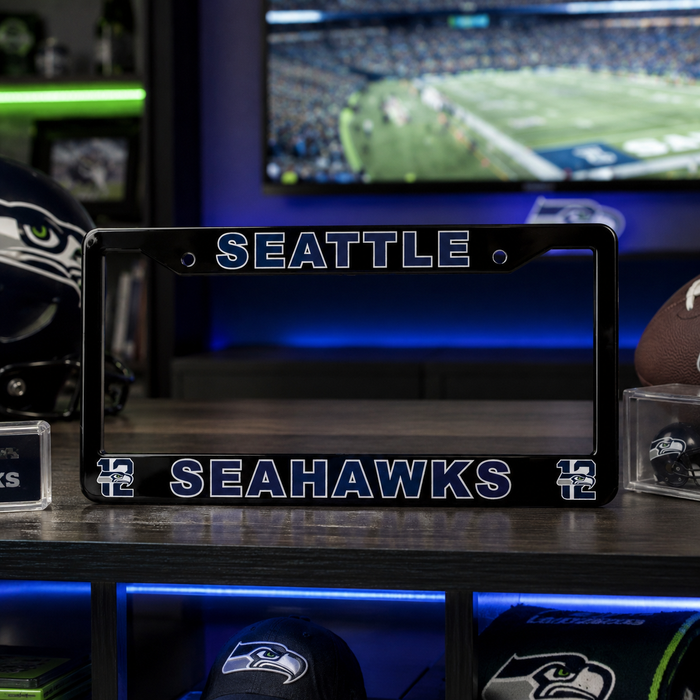 A black license plate frame featuring the Seattle Seahawks logo and team name in blue and white, displayed on a shelf with football helmets and a TV in the background.