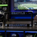 A black license plate frame featuring the Seattle Seahawks logo and team name in blue and white, displayed on a shelf with football helmets and a TV in the background.
