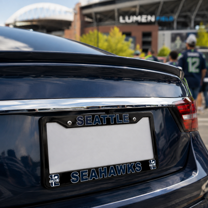 A black license plate frame with 'SEATTLE SEAHAWKS' in blue and white lettering, mounted on the rear of a dark blue car. The background shows a blurred stadium scene with people and a Lumen Field sign.
