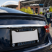 A black license plate frame with 'SEATTLE SEAHAWKS' in blue and white lettering, mounted on the rear of a dark blue car. The background shows a blurred stadium scene with people and a Lumen Field sign.