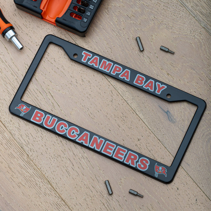 A black license plate frame with 'TAMPA BAY BUCCANEERS' text in red and white, shown on a light wood surface with small mounting screws and a drill bit set nearby.
